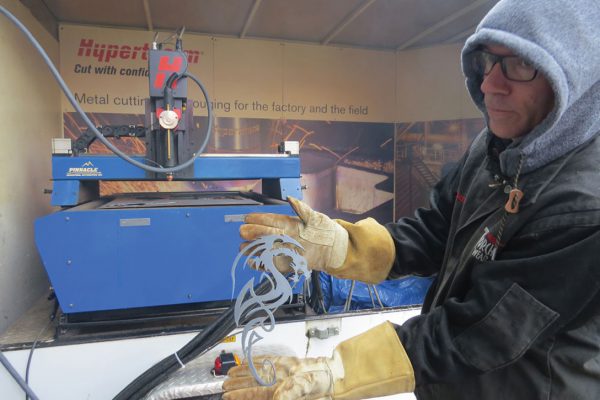 In image of a man in hoodie, jacket and welding gloves holding metal design made with CNC plasma cutter.