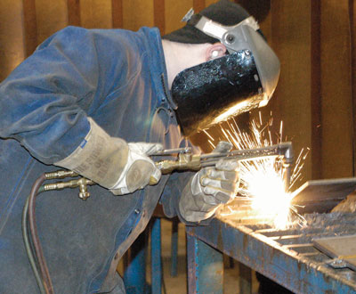 Image shows individual in coveralls and welding mask performing welding tasks, with sparks flying.