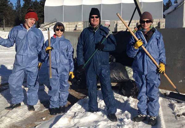 Image of four individuals wearing protective clothing and holding rakes near garbage bags in the snow.