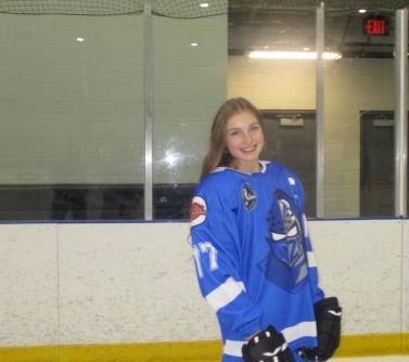 Image shows a young woman with long blonde hair wearing a hockey jersey on the ice.