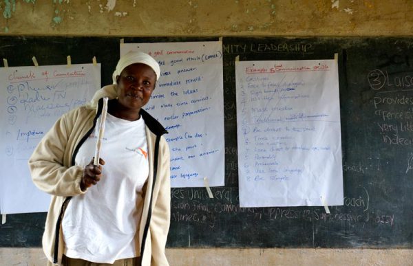 Image shows Kenyan woman standing in front of a chalk board with large pieces of paper with writing taped on it.