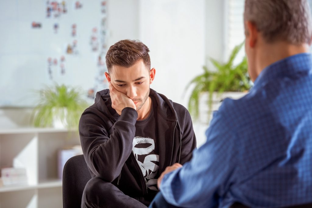 Sad young man discussing with therapist in meeting. Depressed university student talking to mental health professional. They are sitting in lecture hall.