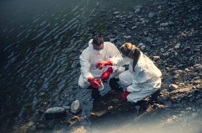 Men and women scientists are looking at a sample of polluted water in the forest.They are wearing protective gloves and clothing, protective face mask