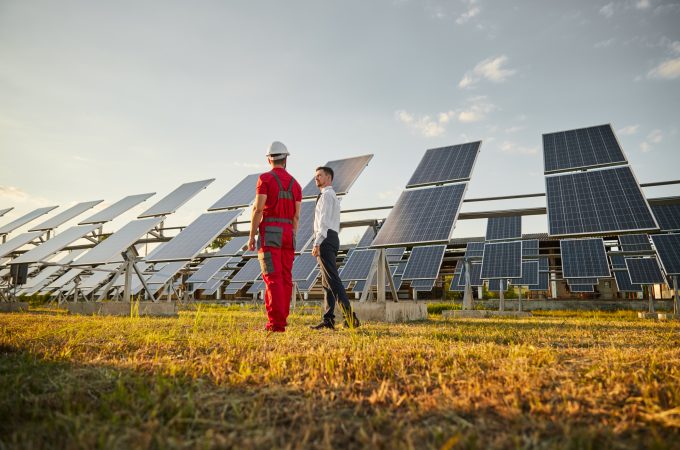 Full body of male investor in formal wear discussing work with engineer in workwear in spacious field with photovoltaic panels working on solar energy and producing eco power