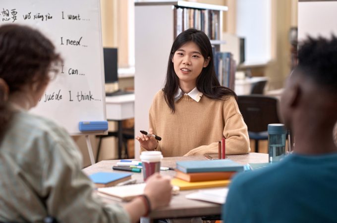 Front view portrait of young Asian woman as female teacher talking to group of students while sitting at table in class