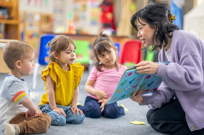 A preschool teacher sits on the floor of her classroom with a small group of students as she reads them a book. The children are each dressed casually and are focused on the story.